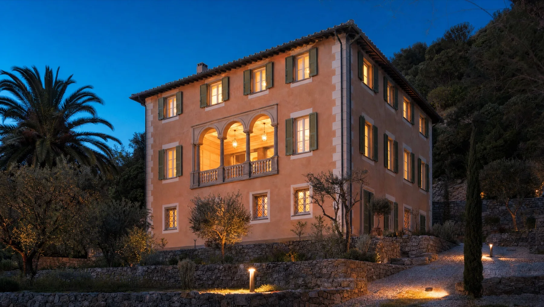 La stessa villa dopo il tramonto, con le finestre illuminate dall'interno e i faretti che segnano il vialetto di ghiaia e i muri a secco